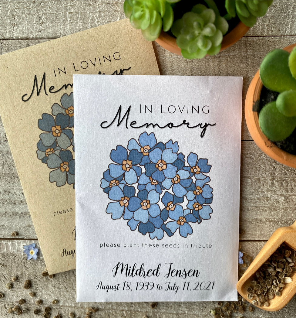 A brown and a white packet with a cluster of blue forget me not flowers and "In Loving Memory."