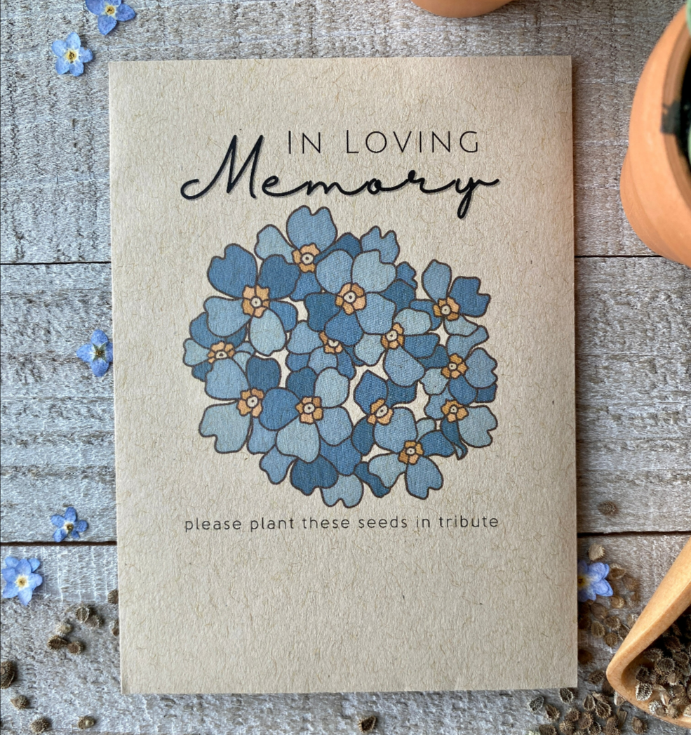 A brown seed packet with a cluster of blue forget me not flowers and "In Loving Memory."